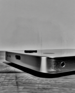 Schwarzweiß-Nahaufnahme der Unterseite und Rückseite eines rahmenlosen Elektronikgeräts aus Metall. Sichtbar sind ein USB-C-Anschluss, zwei kleine Löcher sowie eine Kameralinse auf der Rückfläche. Das Gerät liegt auf einer Holzoberfläche, fotografiert in flachem Winkel von unten.