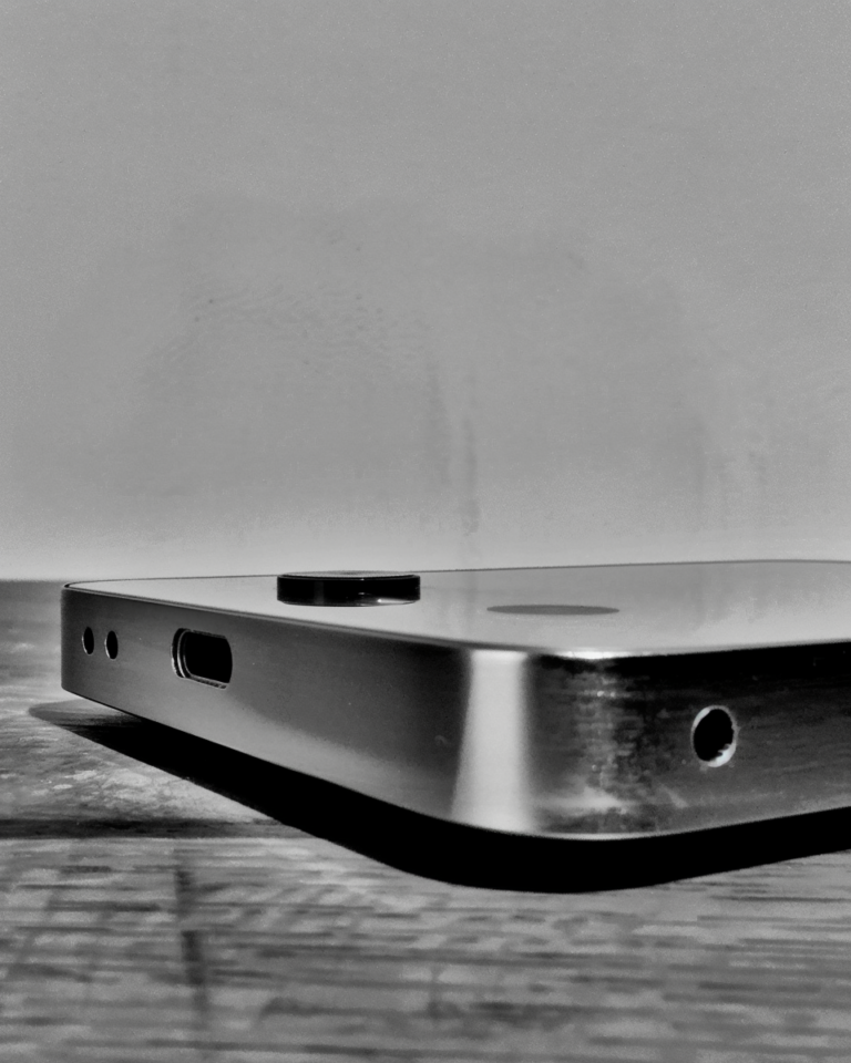 Schwarzweiß-Nahaufnahme der Unterseite und Rückseite eines rahmenlosen Elektronikgeräts aus Metall. Sichtbar sind ein USB-C-Anschluss, zwei kleine Löcher sowie eine Kameralinse auf der Rückfläche. Das Gerät liegt auf einer Holzoberfläche, fotografiert in flachem Winkel von unten.