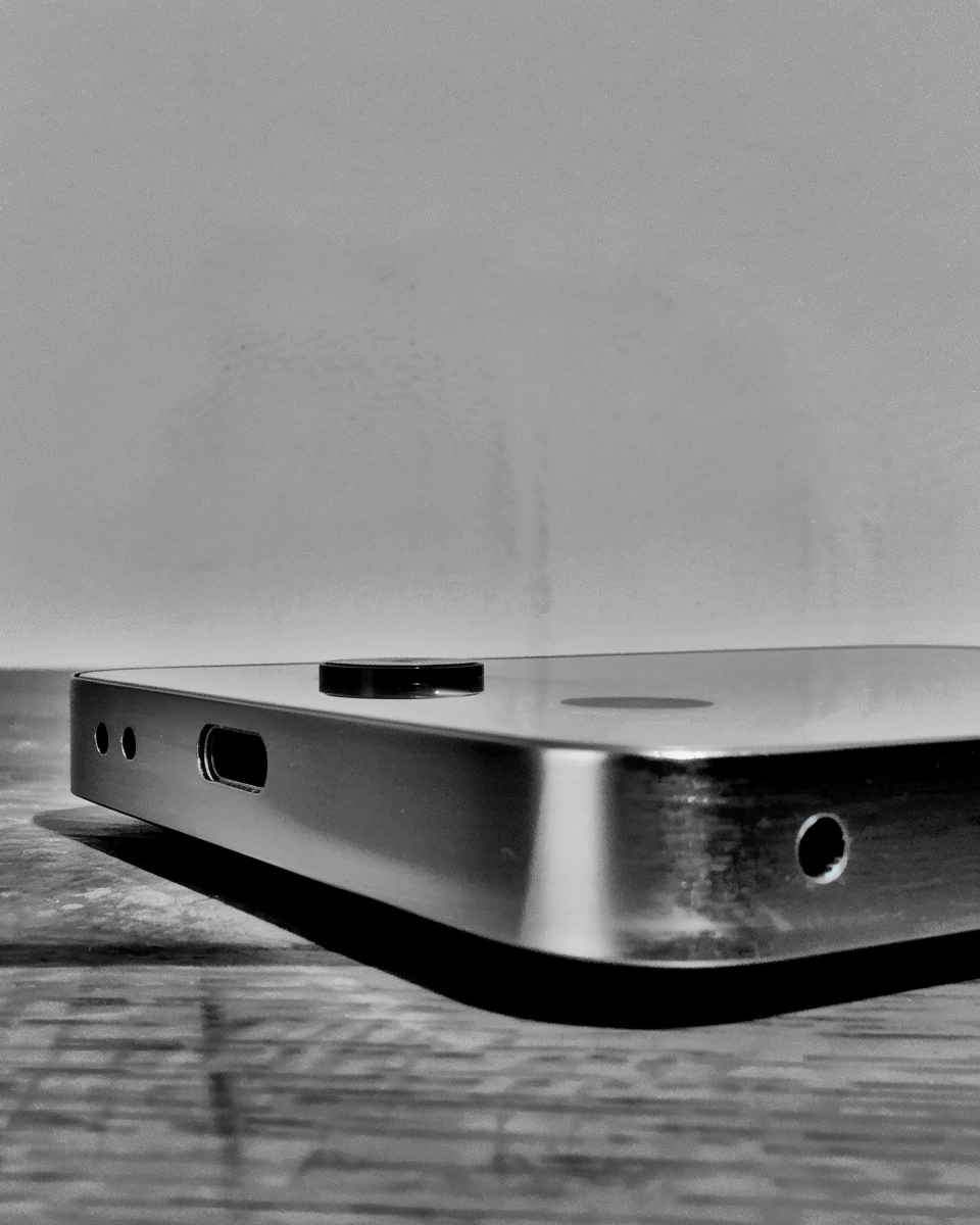 Schwarzweiß-Nahaufnahme der Unterseite und Rückseite eines rahmenlosen Elektronikgeräts aus Metall. Sichtbar sind ein USB-C-Anschluss, zwei kleine Löcher sowie eine Kameralinse auf der Rückfläche. Das Gerät liegt auf einer Holzoberfläche, fotografiert in flachem Winkel von unten.