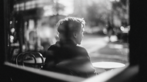 Eine Person sitzt von hinten sichtbar allein an einem Cafétisch am Fenster. Schwarzweißfotografie, unscharfer Straßenhintergrund, Filmkornoptik.