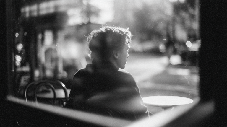 Eine Person sitzt von hinten sichtbar allein an einem Cafétisch am Fenster. Schwarzweißfotografie, unscharfer Straßenhintergrund, Filmkornoptik.